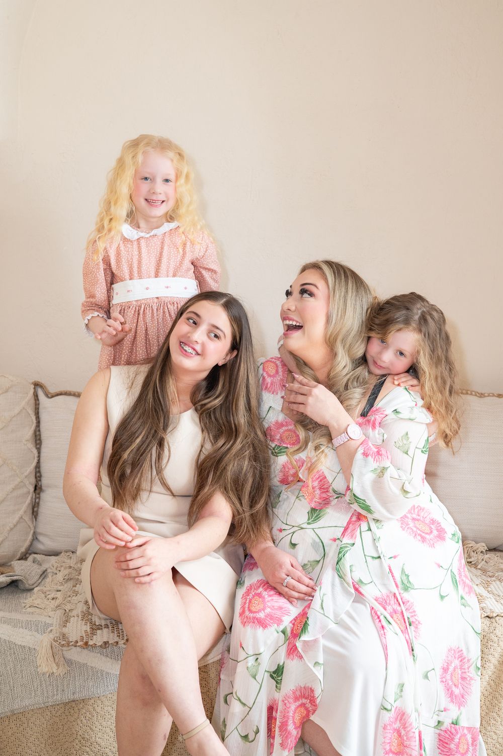 Group of people laughing together while sitting on a beige couch in floral and pink dresses creating a joyful atmosphere.