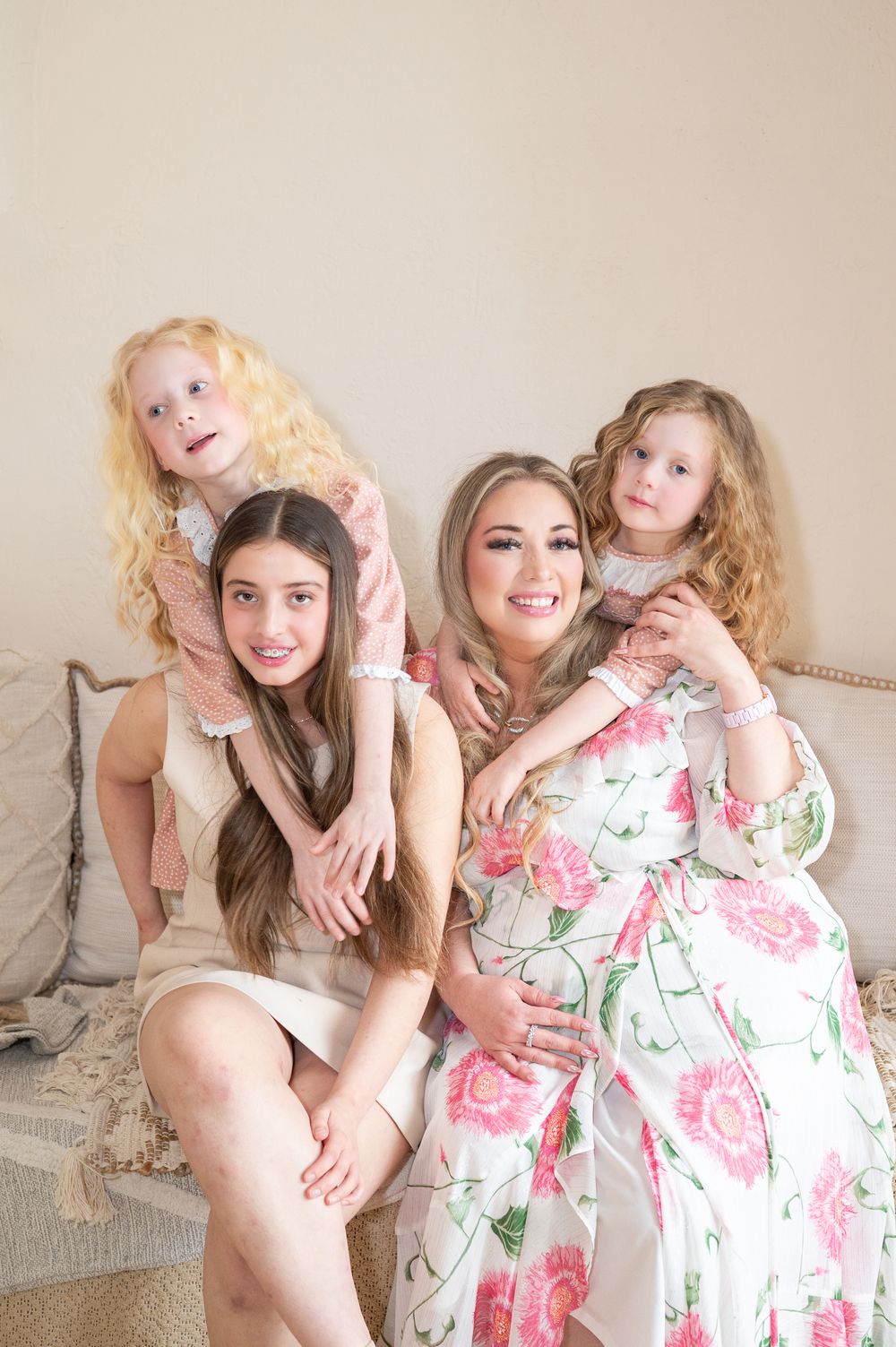 A family group poses together on a beige couch wearing light colored dresses in a soft, warm indoor setting.