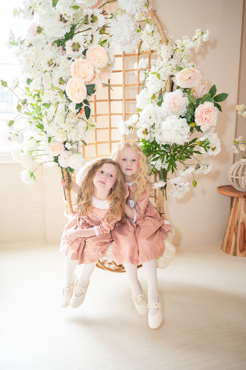 Two in matching pink dresses sit on a decorative swing adorned with blush and white floral arrangements against lattice backdrop.