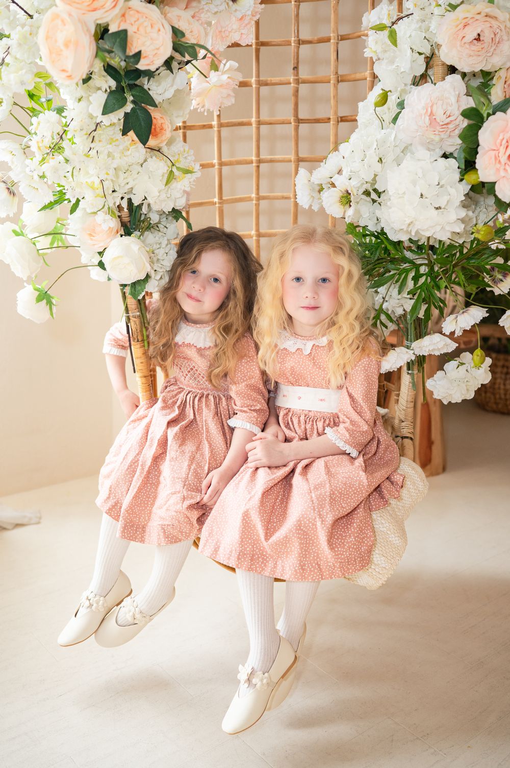 Two young children in matching pink dresses and white stockings pose beneath an elegant floral arch with roses and hydrangeas.