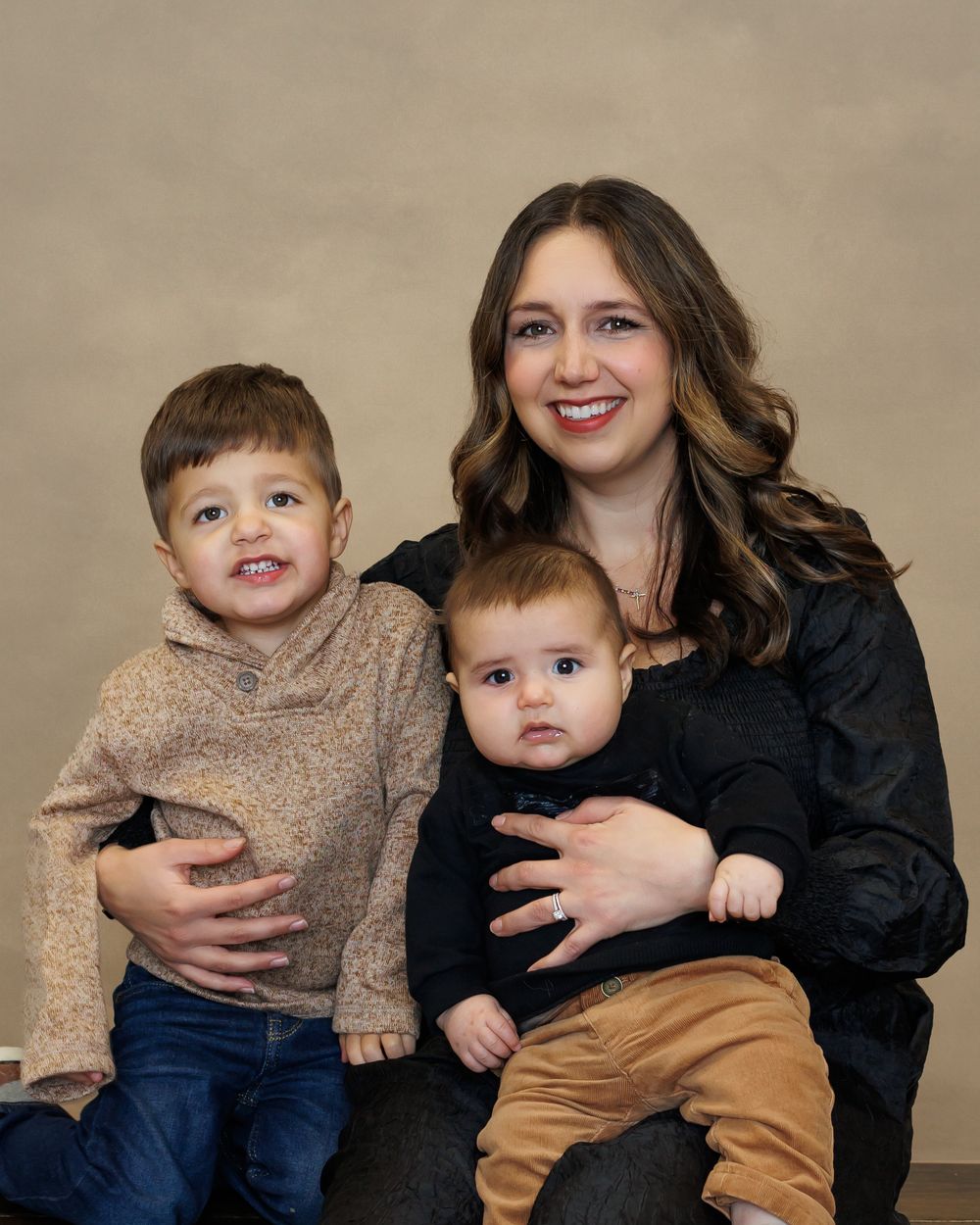 A family portrait with long dark wavy hair in a black top posing with two young children in casual attire.