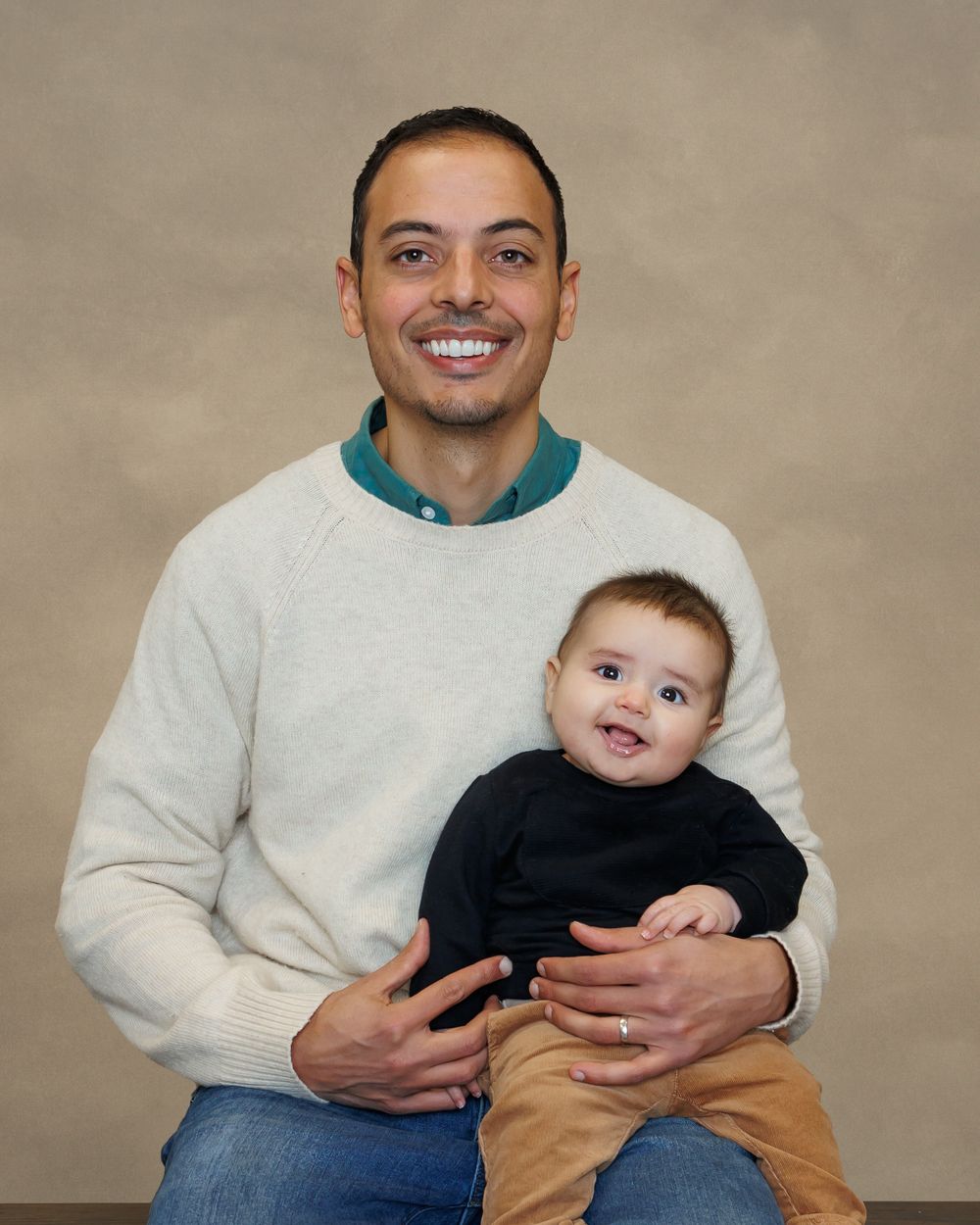 Studio portrait of an adult in a white sweater sitting and holding a happy baby wearing a black shirt and khaki pants.