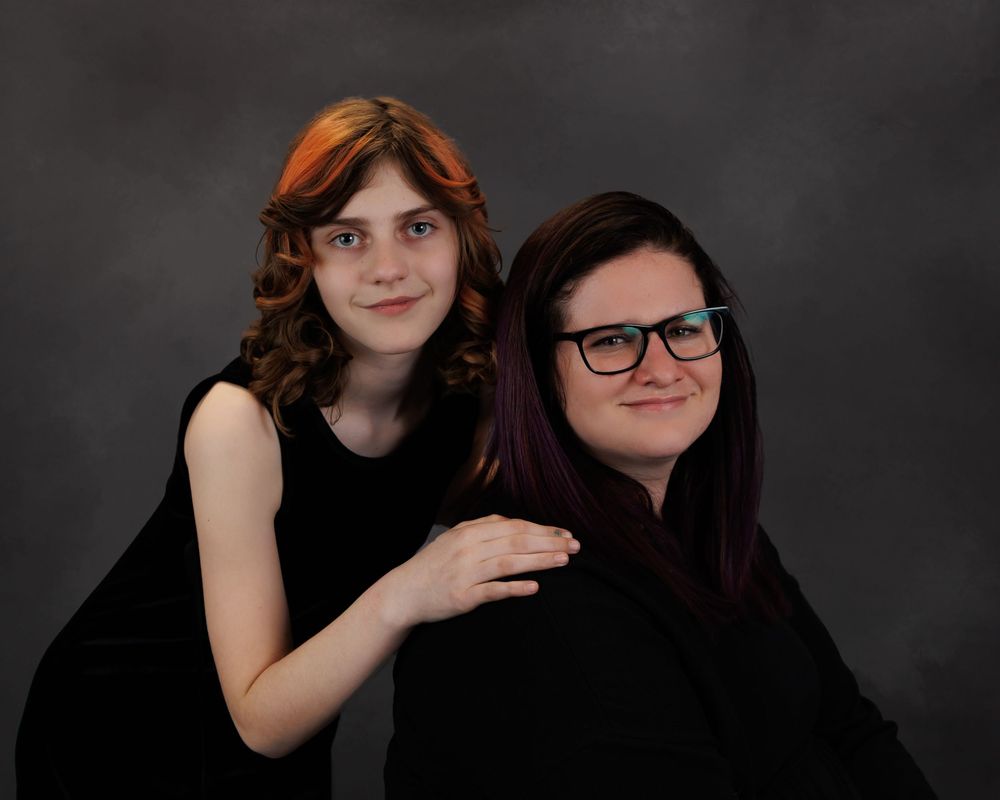 Two people in black attire pose together for a formal portrait against a dark background.