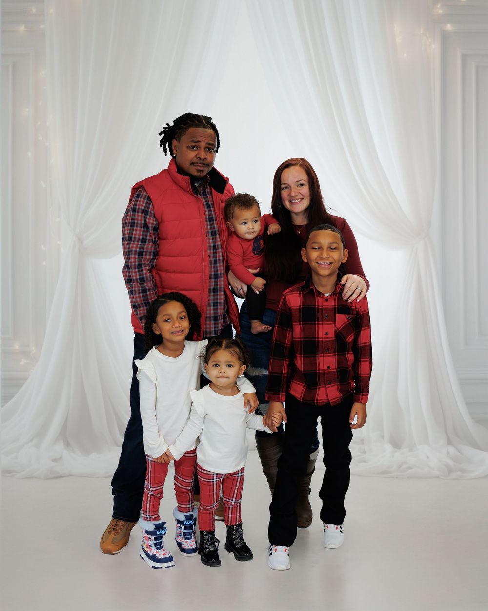 A family of six wearing coordinated red and plaid outfits poses together against a white backdrop.
