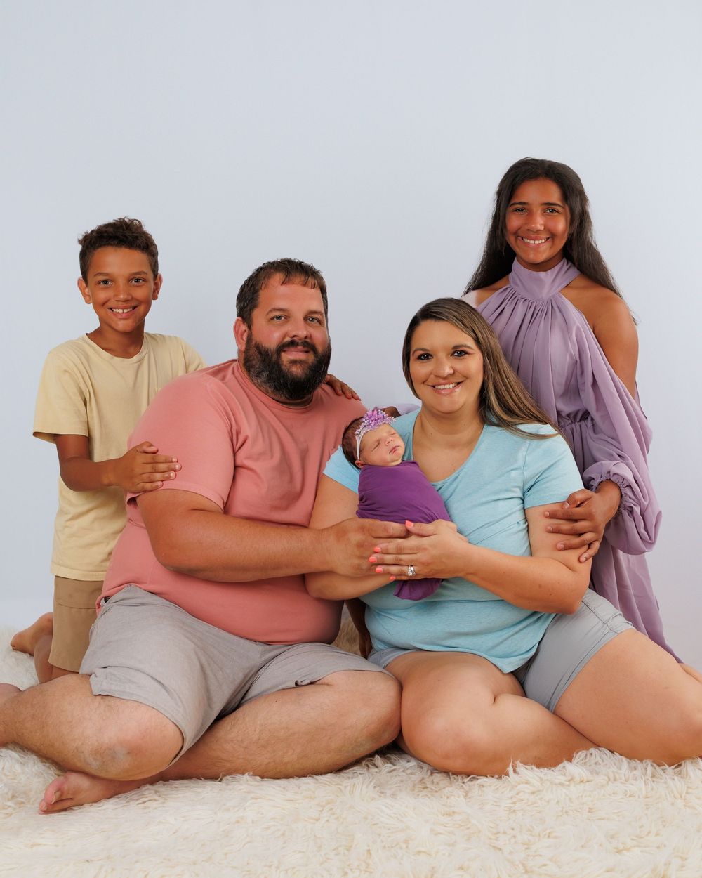 A family of five sitting together on a fluffy white carpet, holding and snuggling with their new baby.