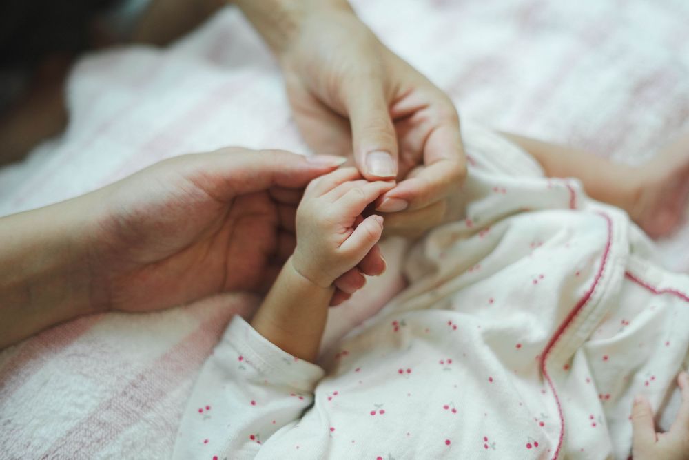 A tiny baby's hand grasps an adult's finger while lying on pink floral bedding.
