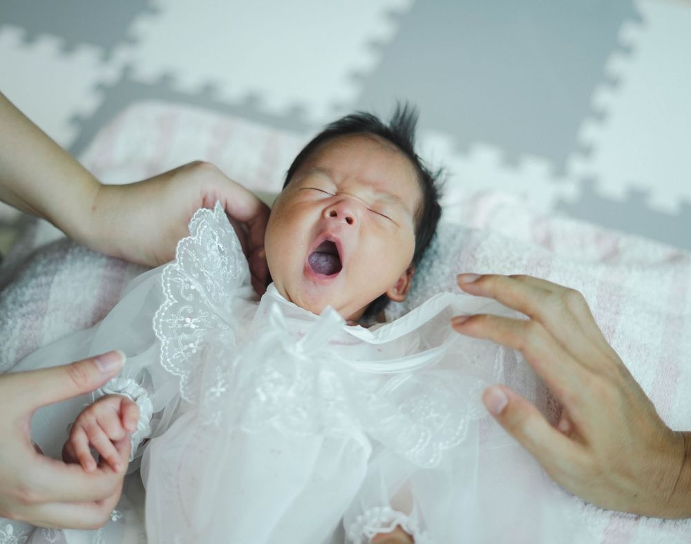 Baby in white lacy dress yawns while lying on soft gray play mat with hands visible supporting them.