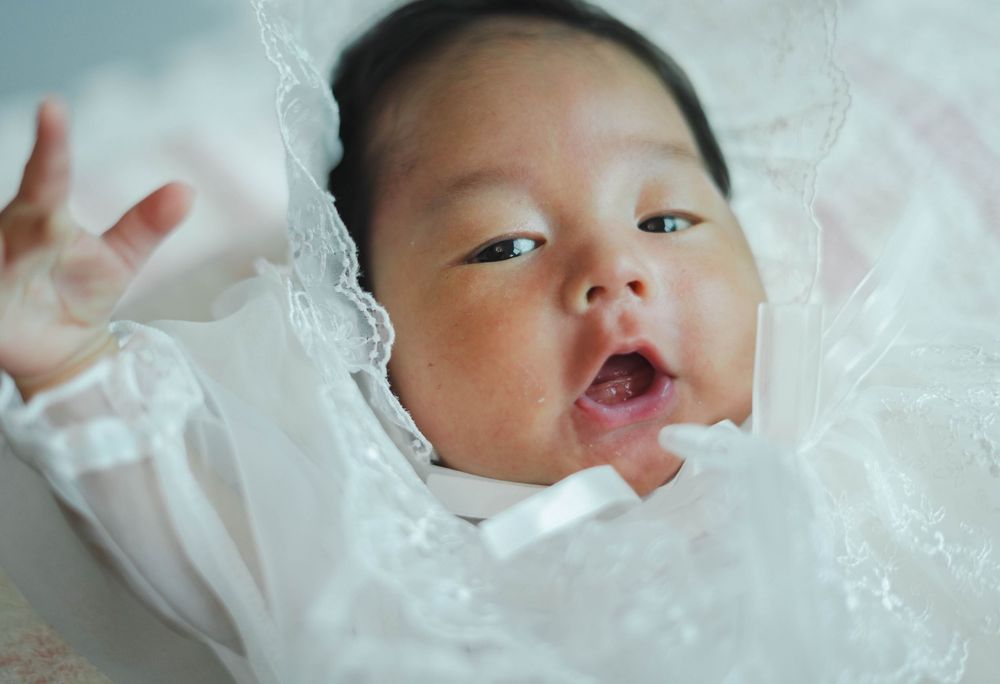 Baby in white lacy outfit smiles joyfully while looking up with bright expression.