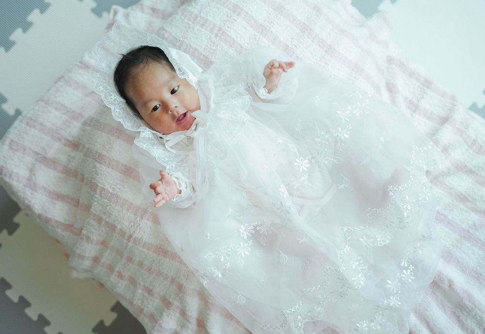 A baby lies on a pink and white striped blanket wearing an ornate white lace christening gown.