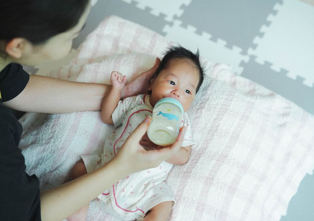 A bottle-feeding moment on a soft play mat with gray foam tiles and a pink blanket.
