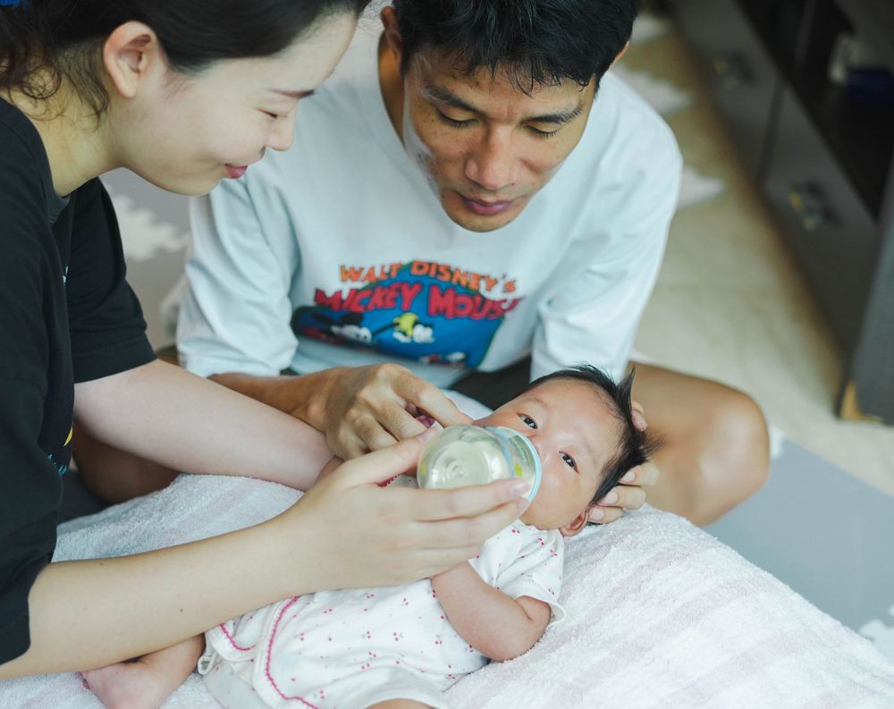 Parents feeding their infant with a bottle while sitting together on the floor.