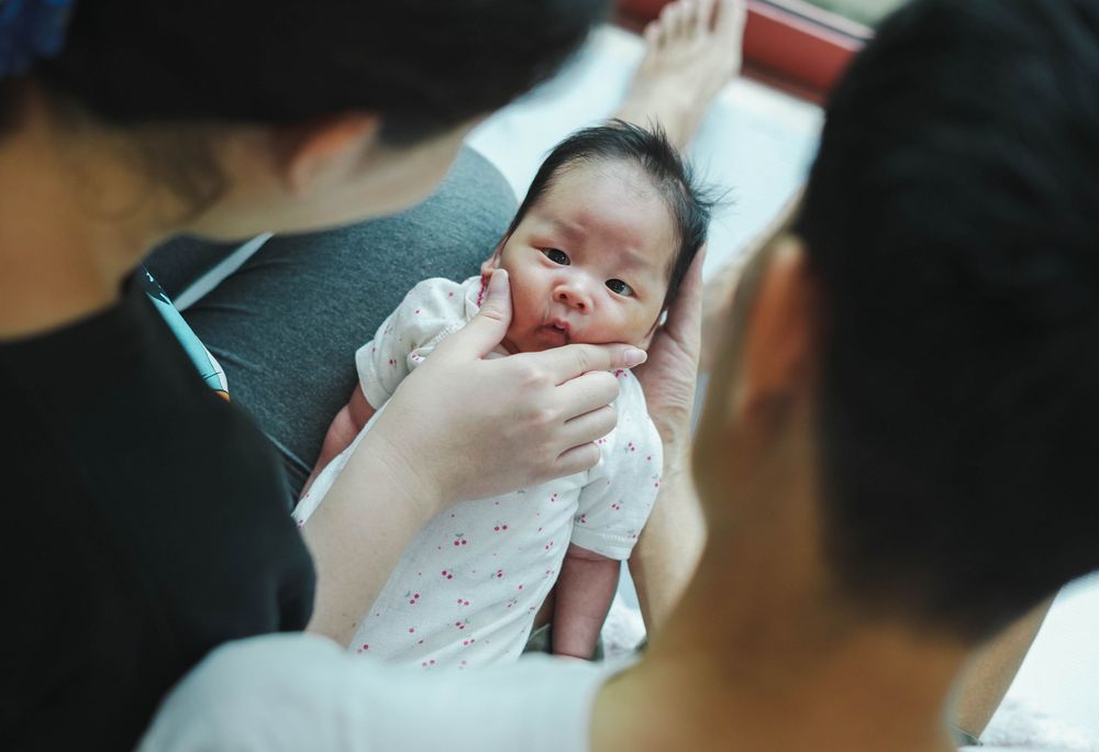 A small newborn baby in white clothing is being gently held and examined by caregivers.