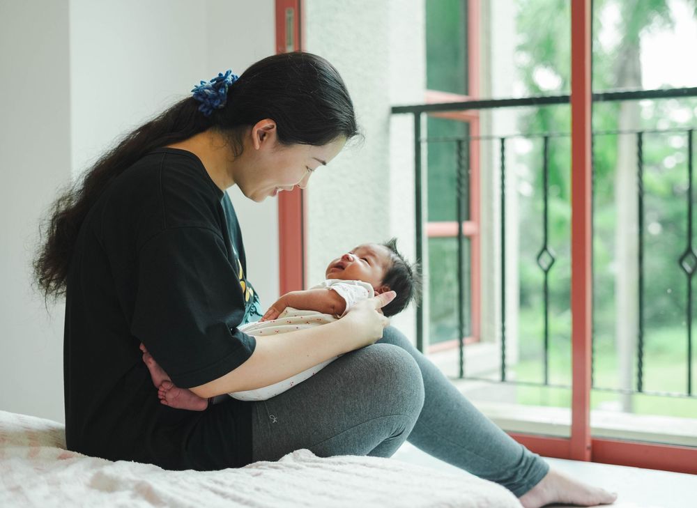 A loving moment between mother and infant sitting on bed by window with natural light streaming in.