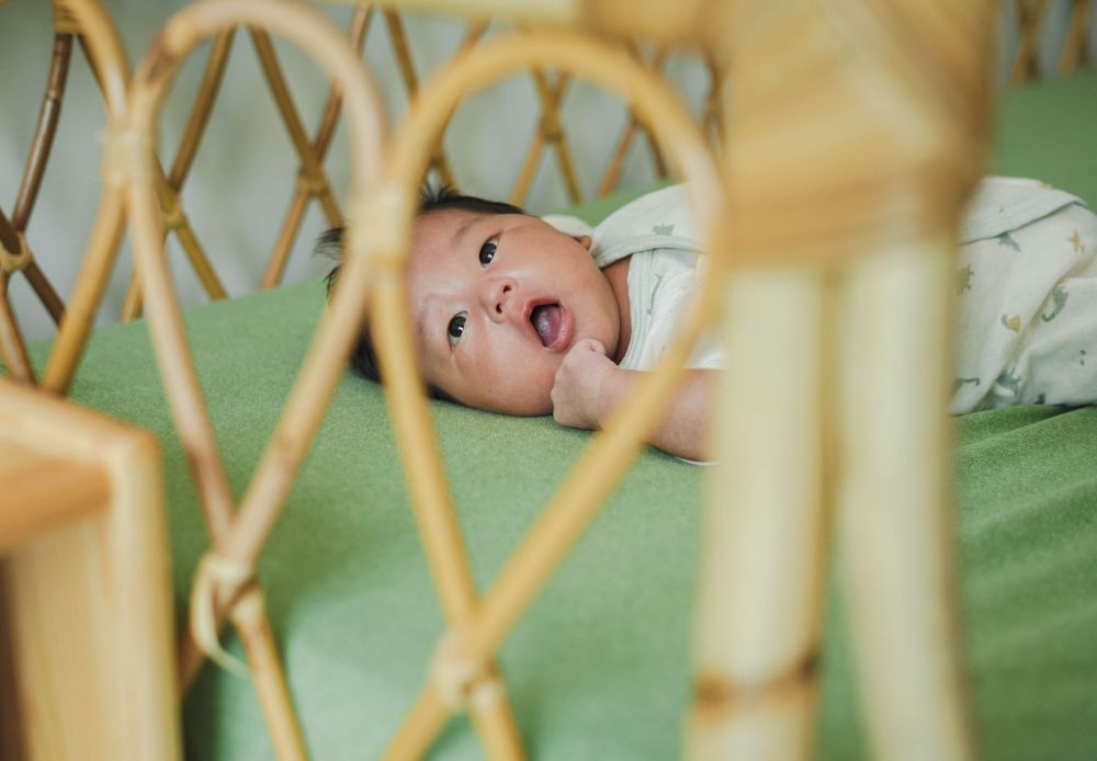 A baby lies on green bedding viewed through wooden crib slats, looking up with an expressive face.