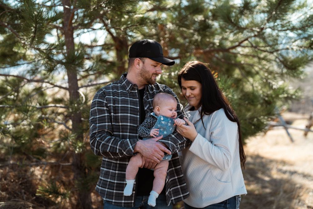 A family stands together in a pine forest on a sunny day enjoying time with their baby.