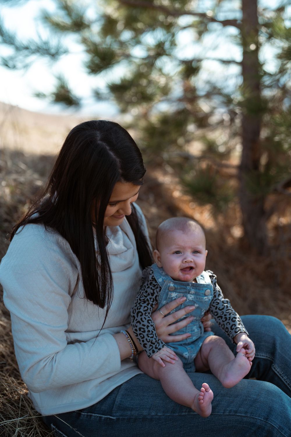 A parent in a white sweater sits outdoors with a smiling baby wearing a patterned outfit against a blurred forest backdrop.