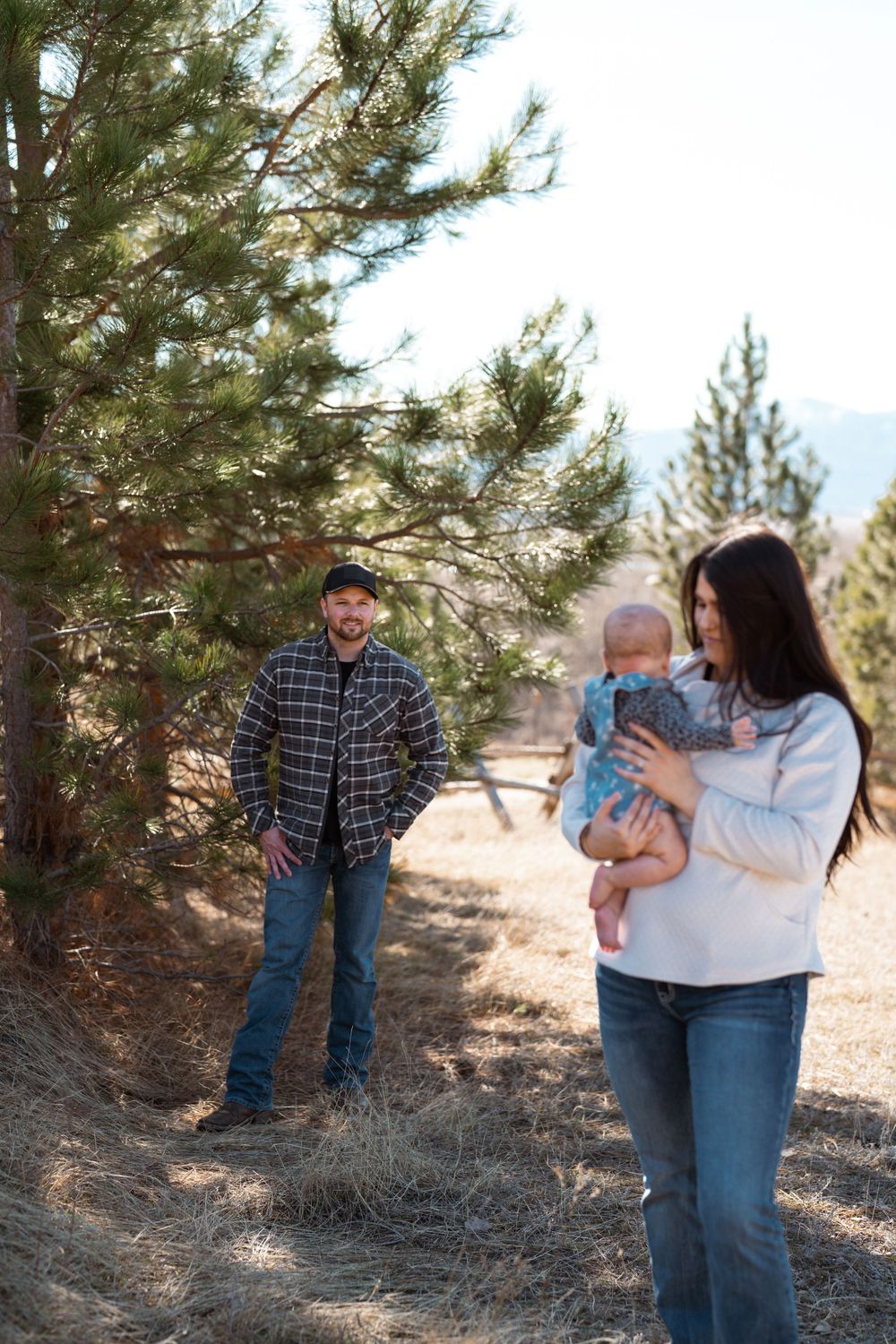 A couple and their baby enjoy a walk among tall pine trees on a sunny mountain trail.