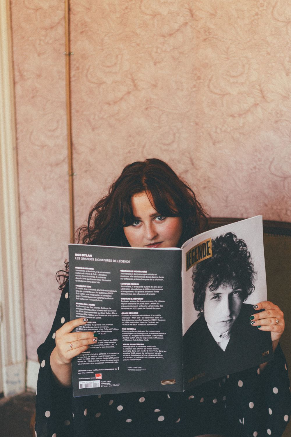 Someone holds up a Bob Dylan album or book while sitting against a textured pink wall.