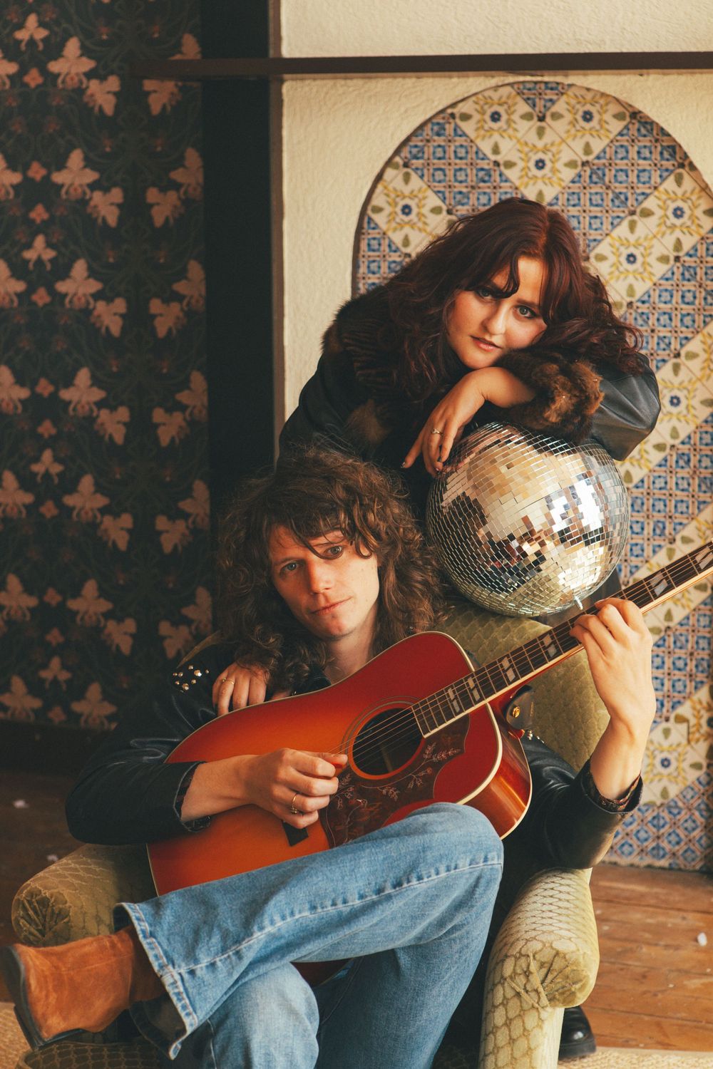 A musician sits on a chair playing a red acoustic guitar against a wall with decorative tiles and patterns.
