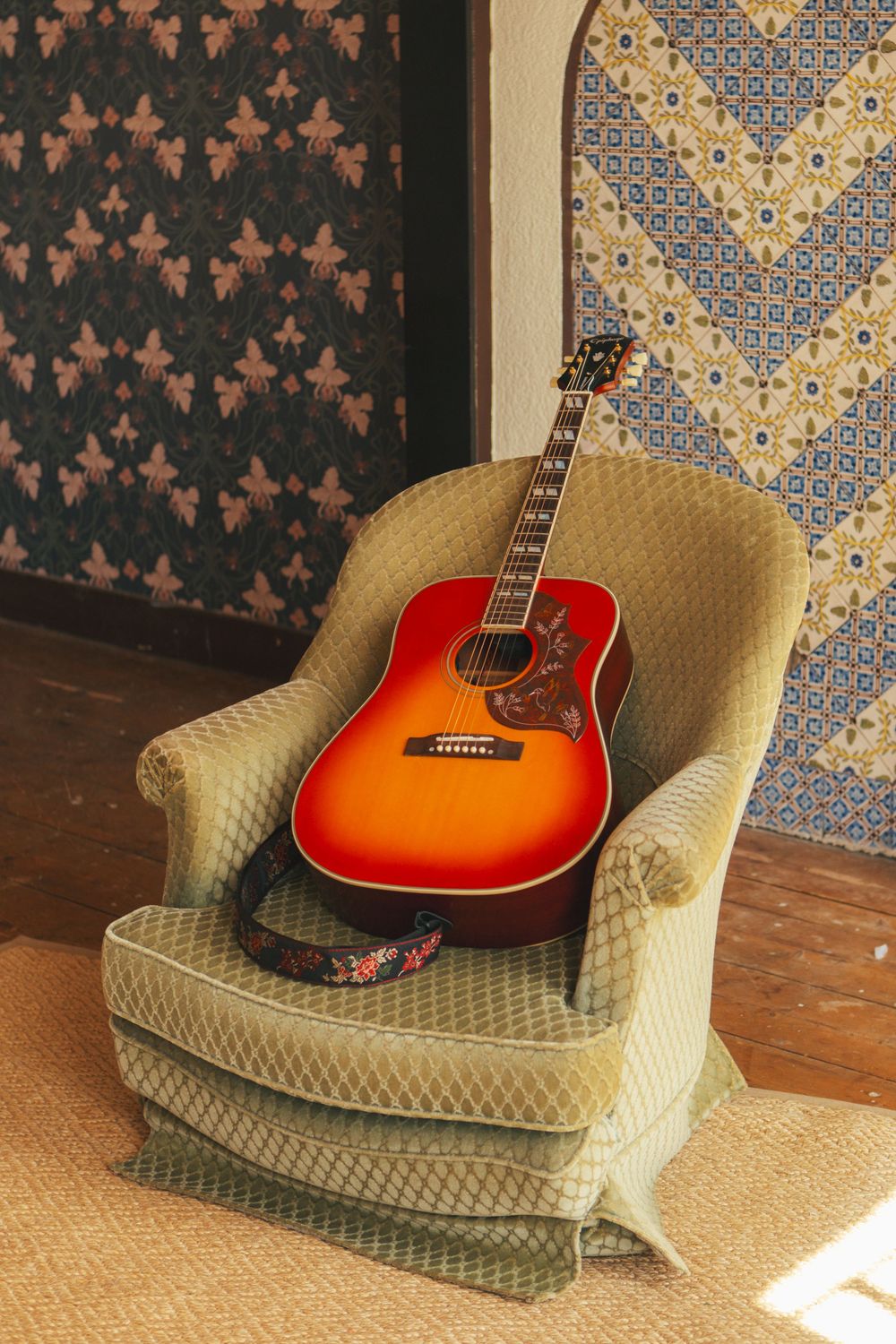 A sunburst acoustic guitar rests on a vintage beige upholstered armchair against a wall with patterned wallpaper.