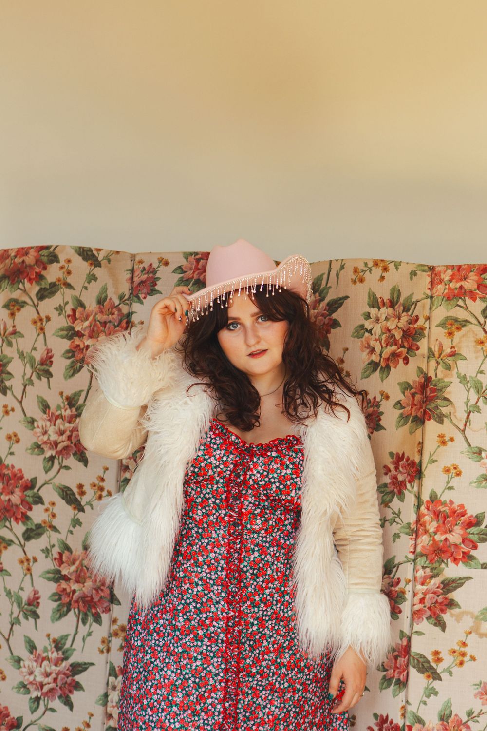 Floral dress layered with fluffy white jacket and pink hat against wallpapered backdrop.