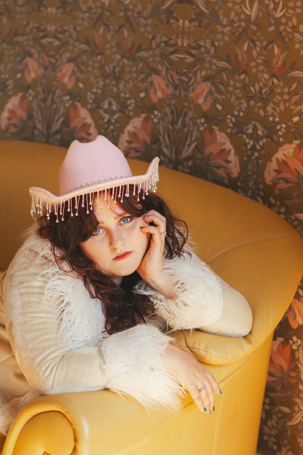 Someone wearing a pink crown with fringe trim reclines on a yellow chair against a floral wallpaper background.