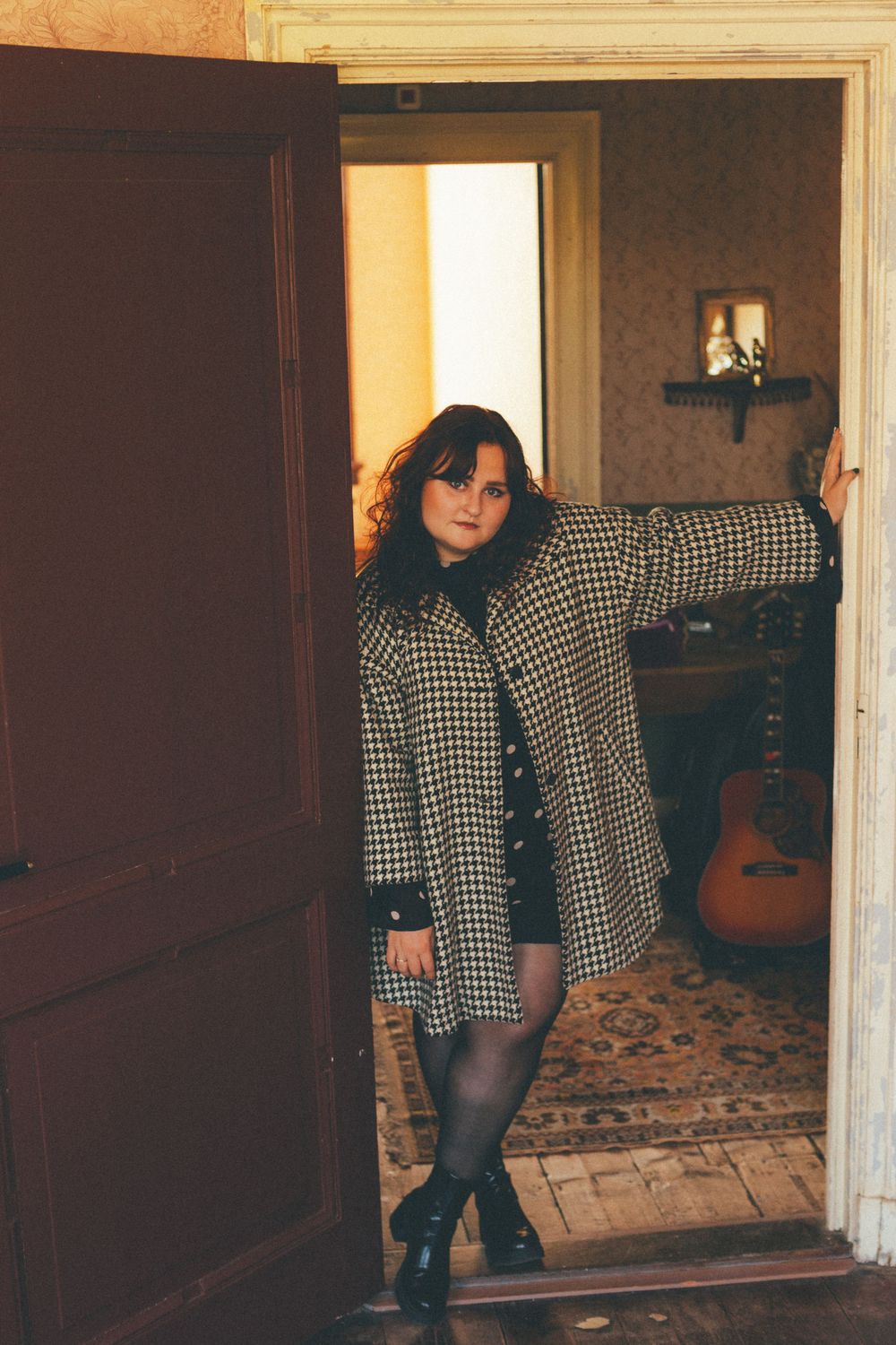 Someone poses in a doorway wearing a black and white checkered coat with black tights and boots in vintage-style setting.