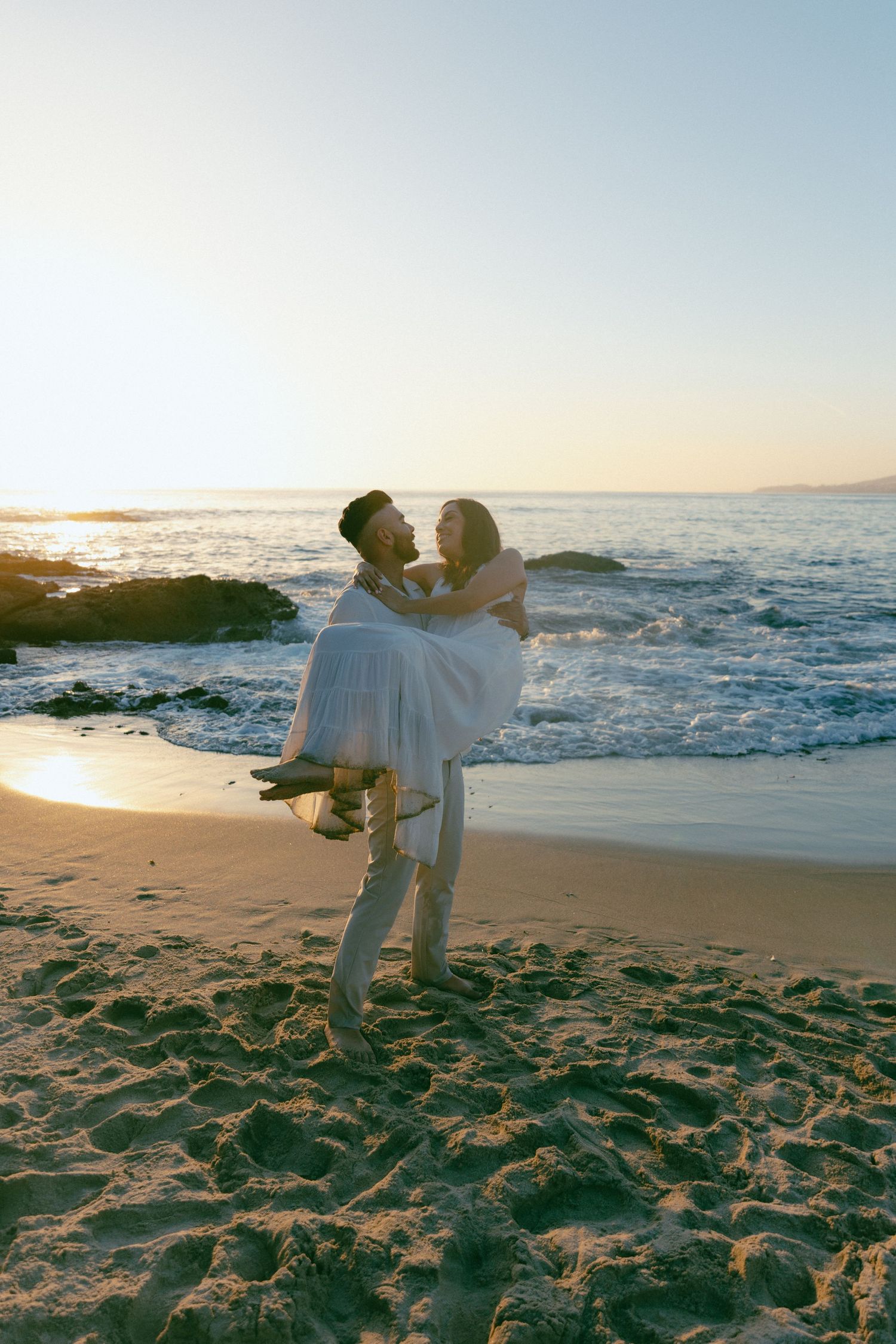 Couple sharing a romantic moment on the beach during sunset with crashing waves and rocky coastline in the background.