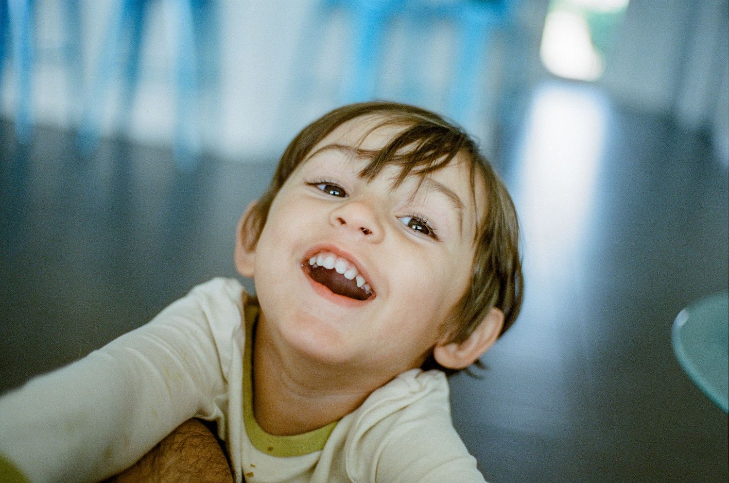 A young child wearing a light yellow shirt smiles warmly with spontaneous joy while looking upward at the camera.