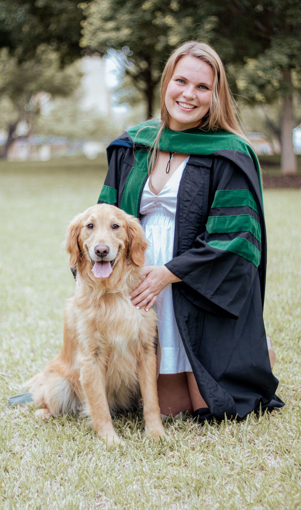 Graduate in black and green academic regalia poses outdoors with friendly Golden Retriever dog on grass.