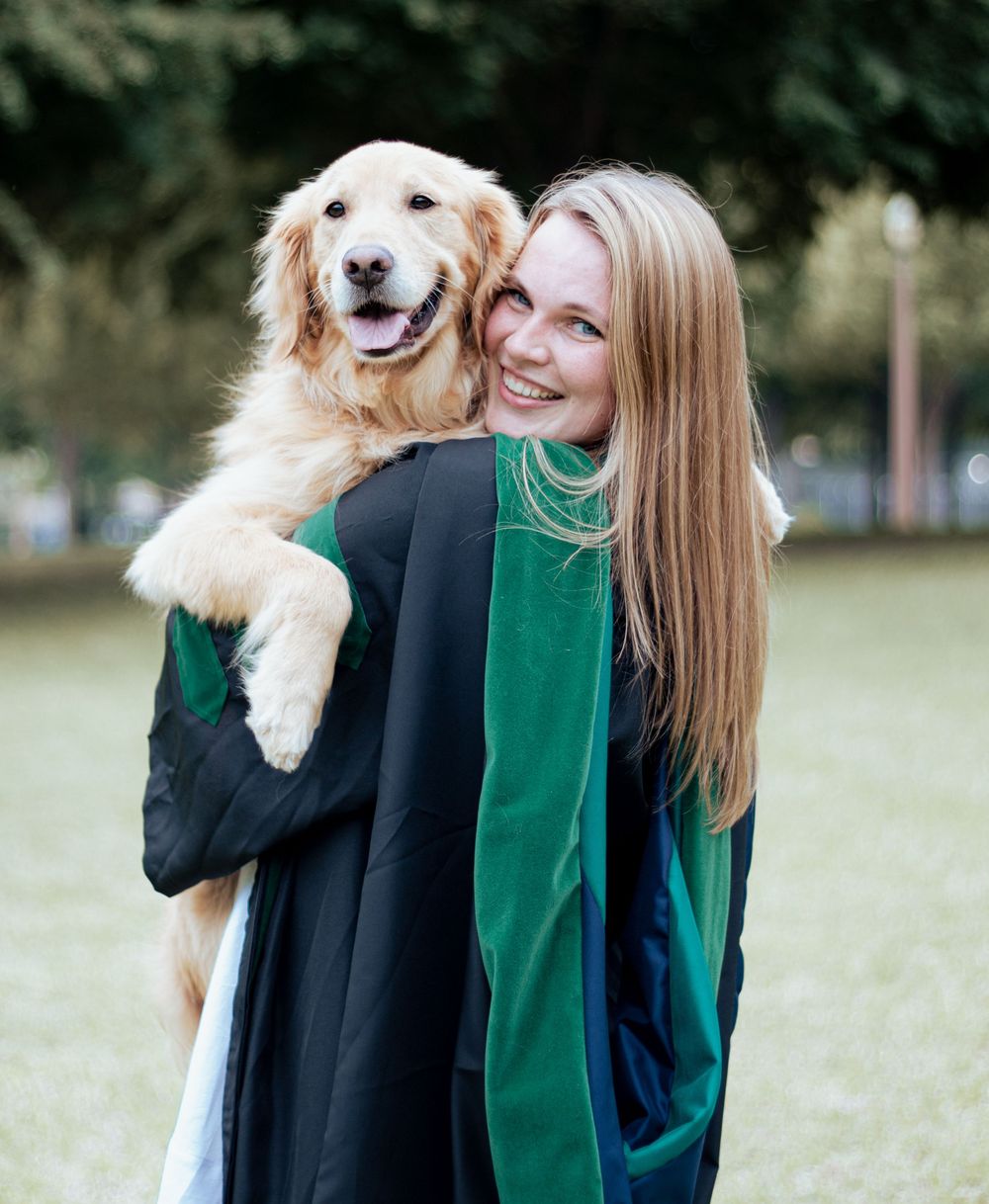 A happy Golden Retriever being held up by someone wearing graduation regalia in green outdoors.