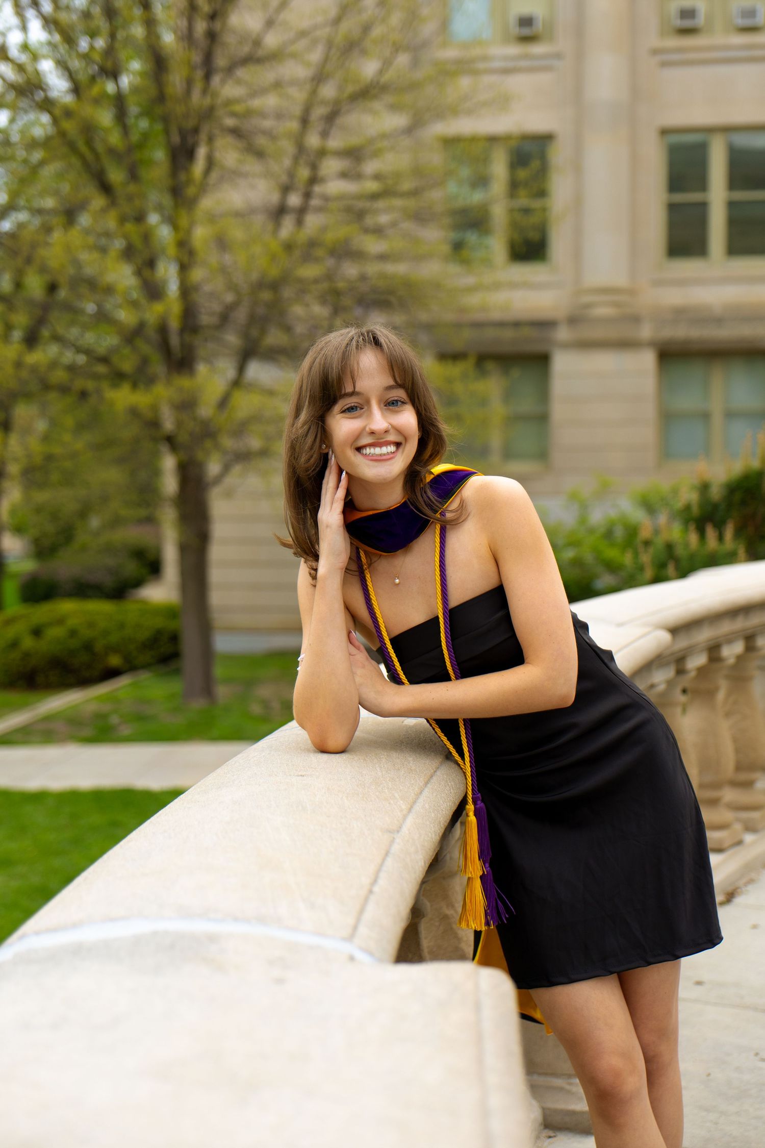 A portrait of a woman in a black dress and graduation regalia leading on a stone ledge with trees in the background.