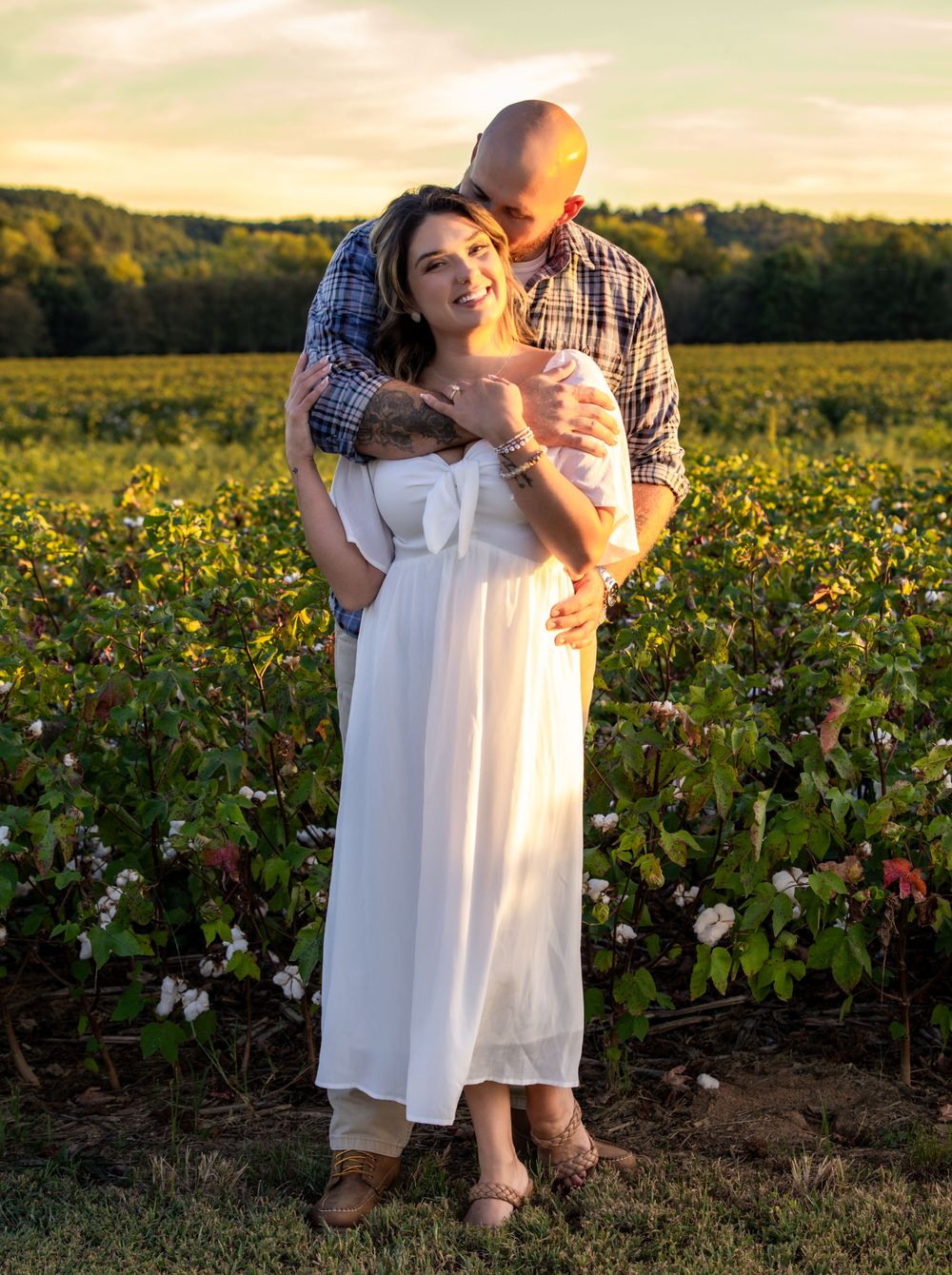 Couple embracing at sunset in lush green field, woman in white dress smiling as man kisses her cheek.