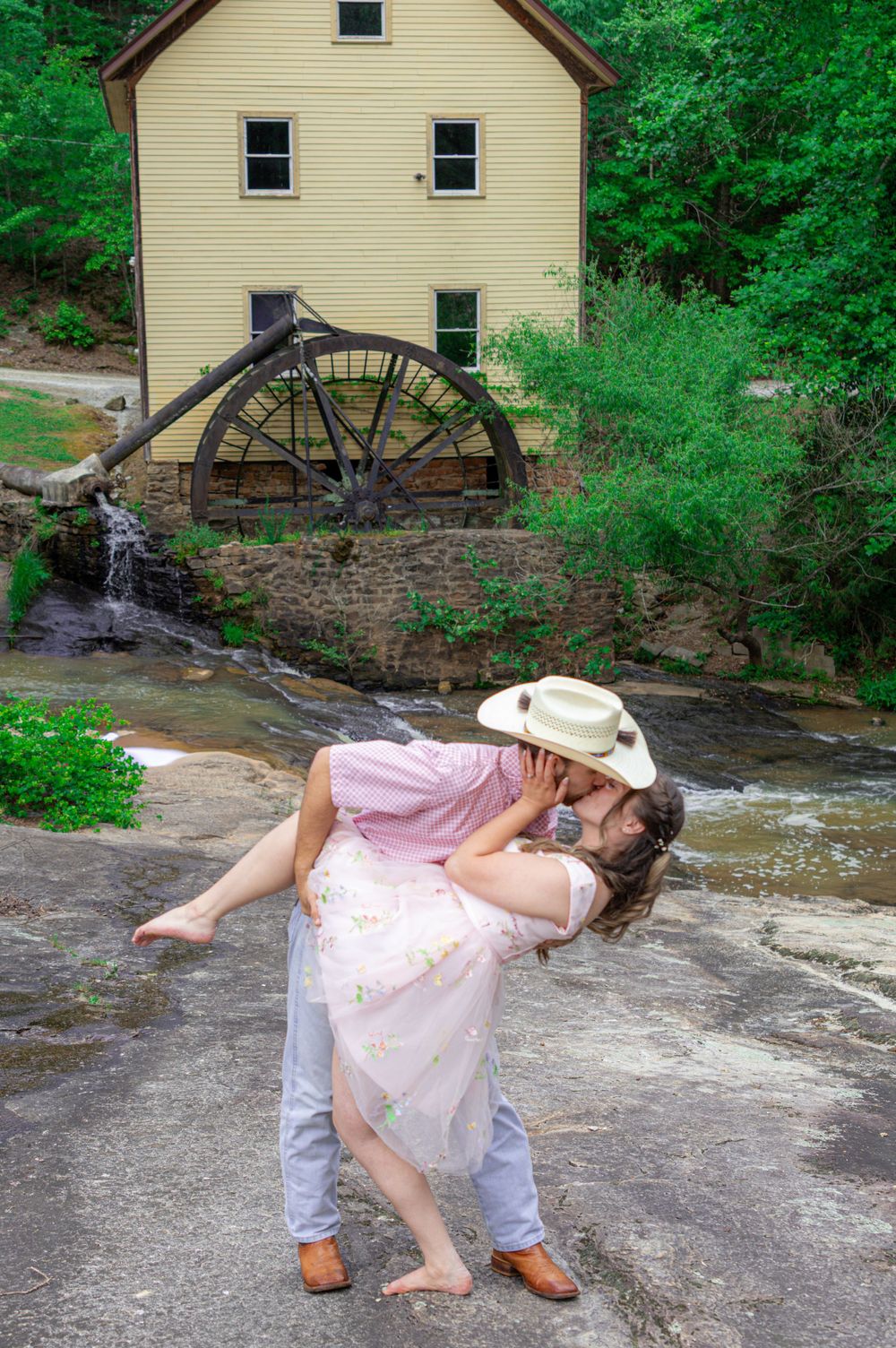 Romantic couple sharing a kiss near a historic grist mill waterwheel, man lifting woman in pink dress outdoors.
