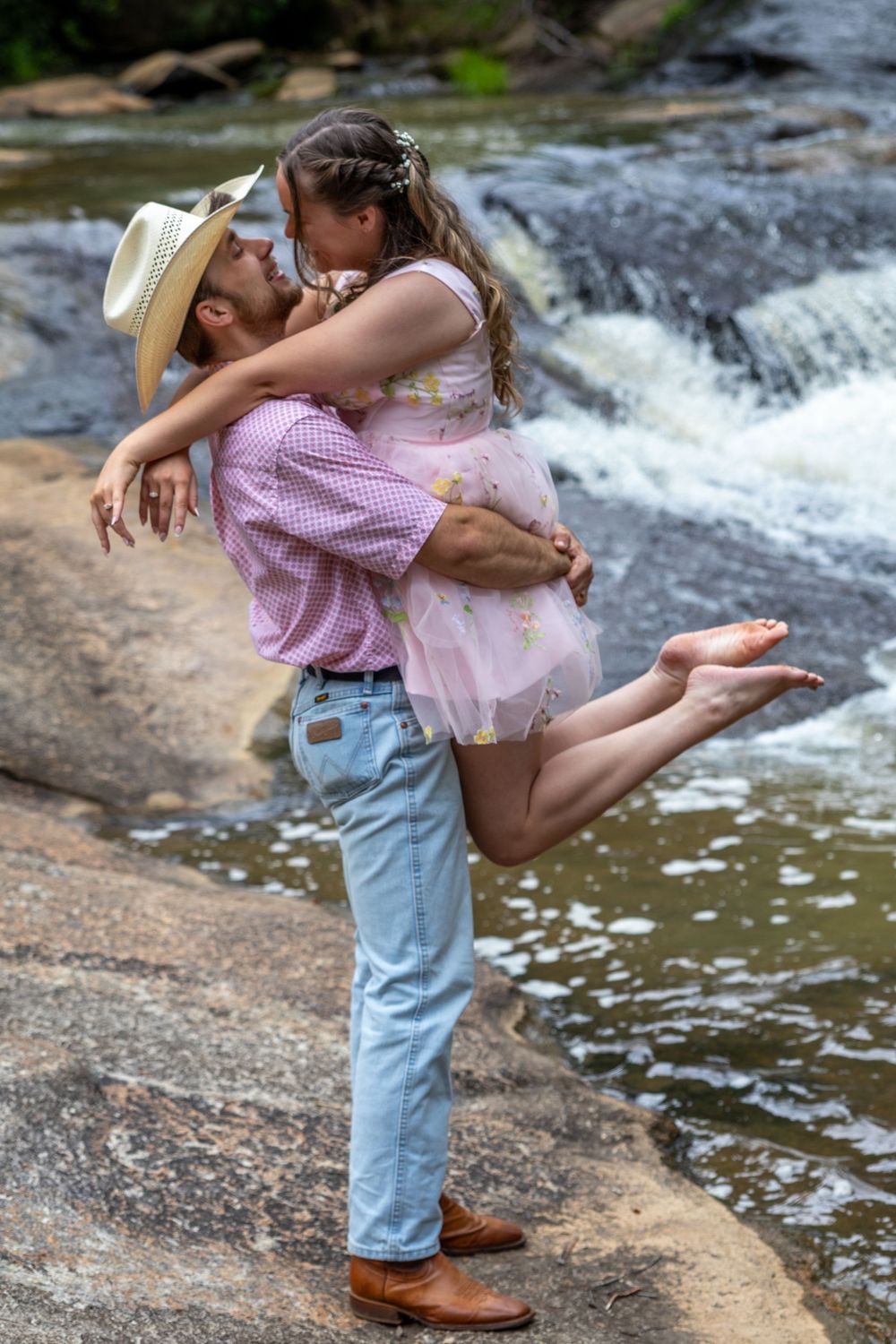 Romantic couple kissing near a waterfall, man lifting barefoot woman in pink dress by rocky stream.