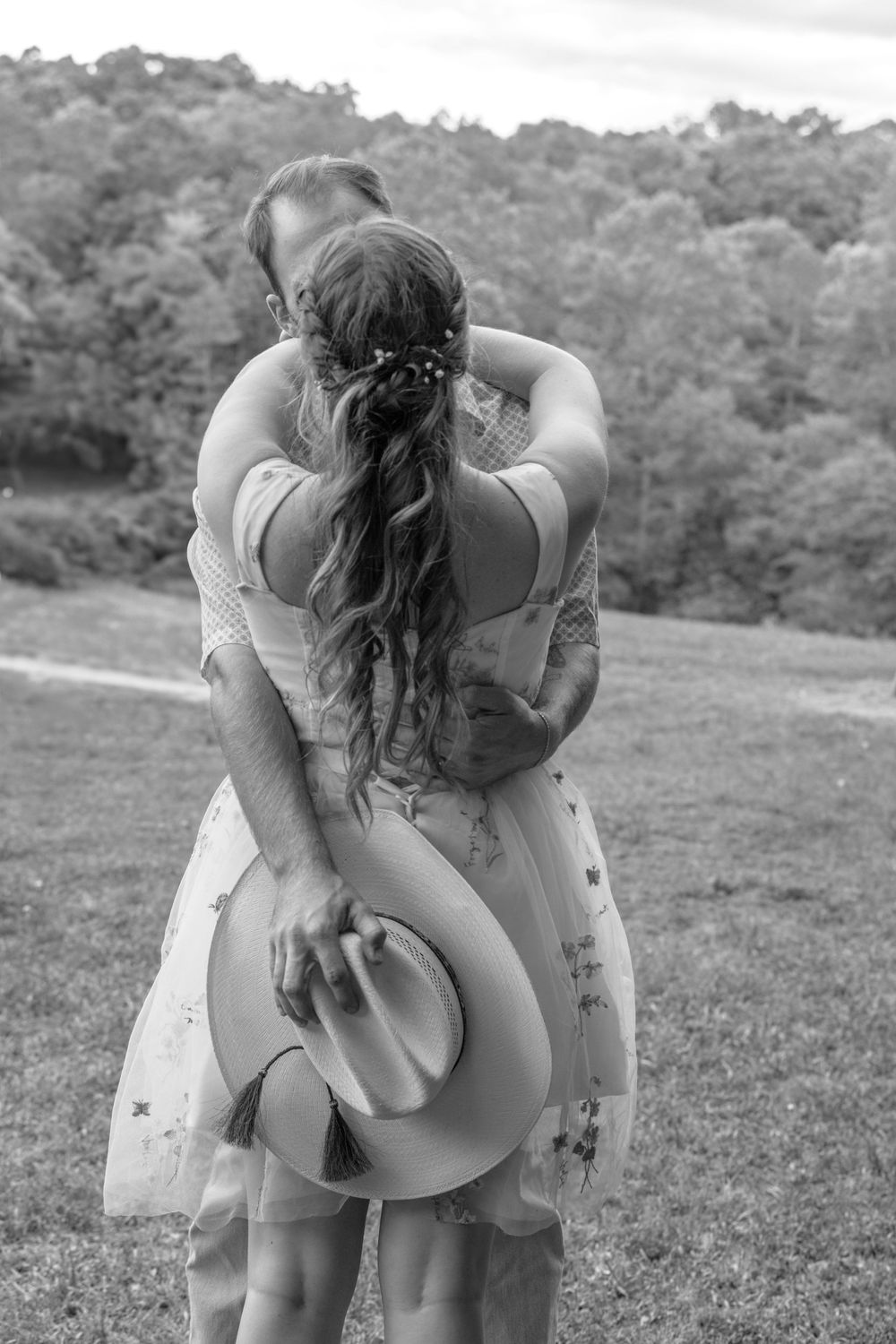 Black and white photo of a couple embracing in a field, man holding cowboy hat behind womans back.