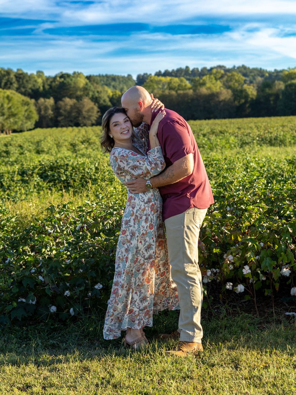 Happy couple hugging in cotton field on sunny day, woman in floral dress and man in burgundy shirt.