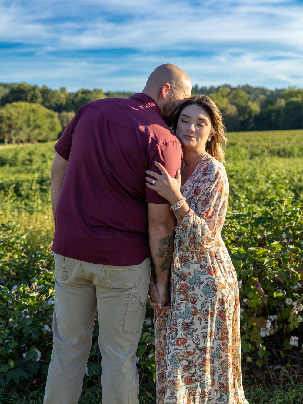 Couple embracing in a lush green field under a blue sky, woman in floral dress smiling warmly.