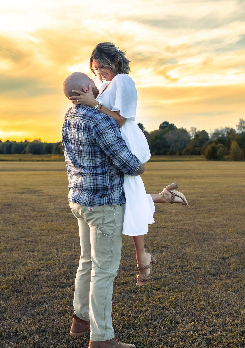 Couple sharing romantic sunset kiss in open field, man lifting woman in white dress during golden hour photography session.