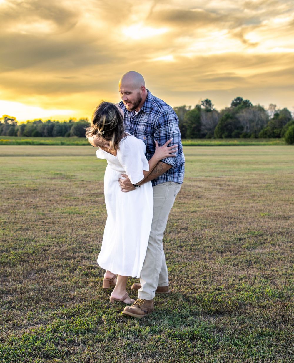Couple embracing in open field at golden sunset, woman in white dress, man in plaid shirt, romantic outdoor photo.