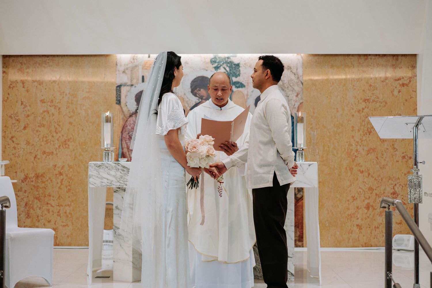 A wedding ceremony in progress with a couple and officiant exchanging vows against a beige wall backdrop.