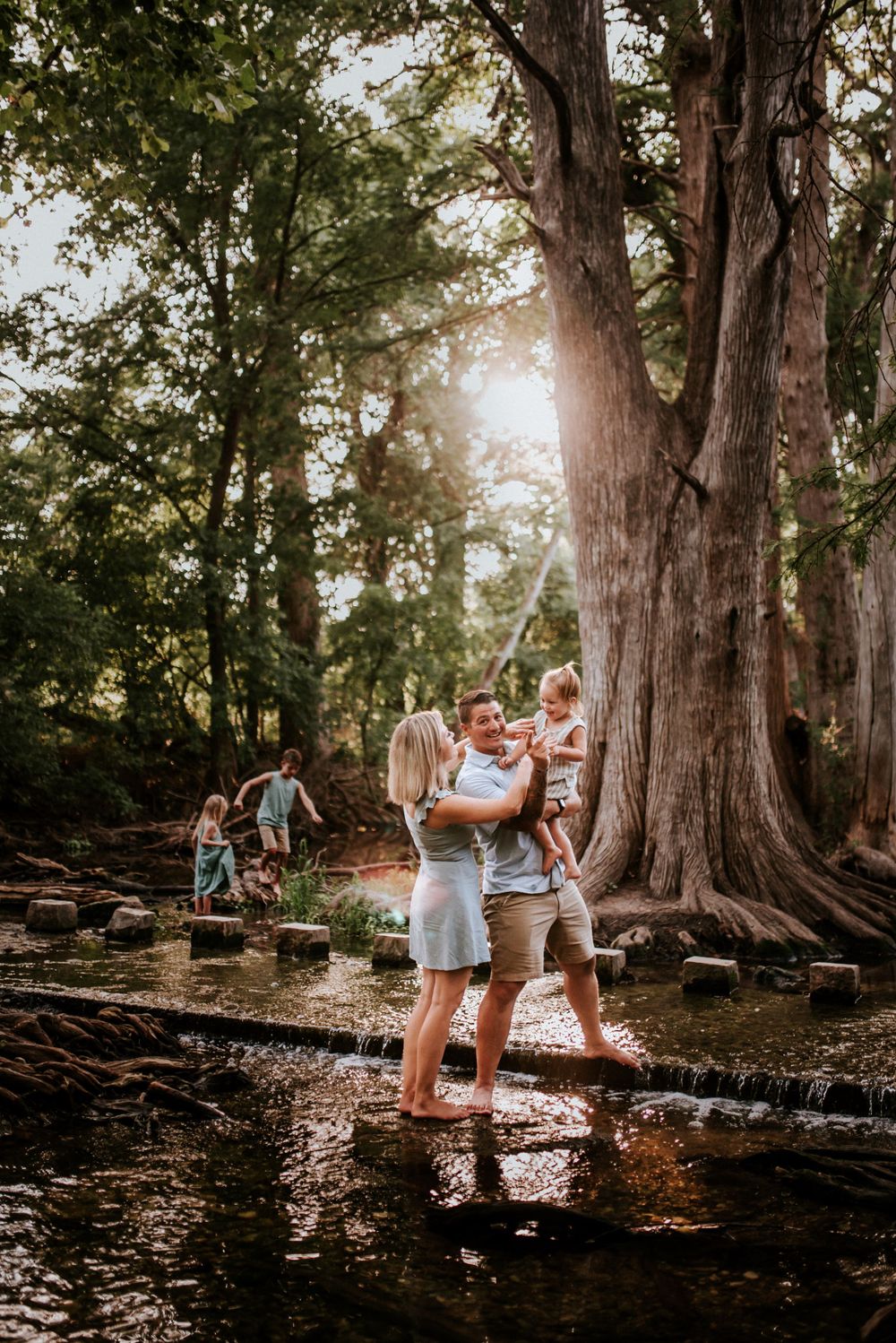 Adventure family session as mother and father hold the youngest while the other children explore the river in Boerne Texas.