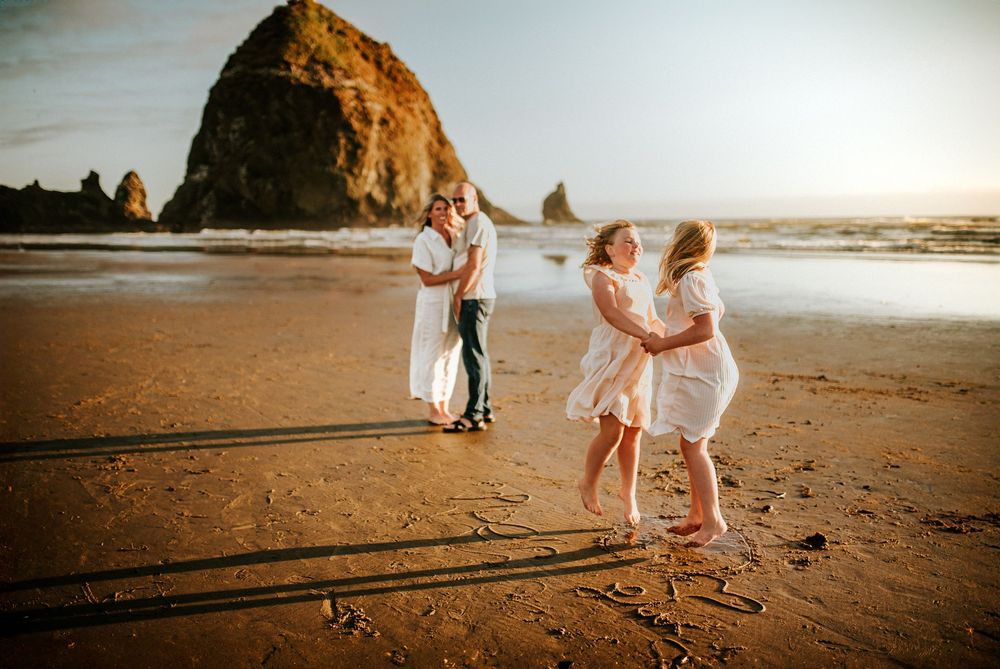 Coastal family at the beach as sisters jump around with happiness together as their parents stand in the background at Canon Beach, Oregon.