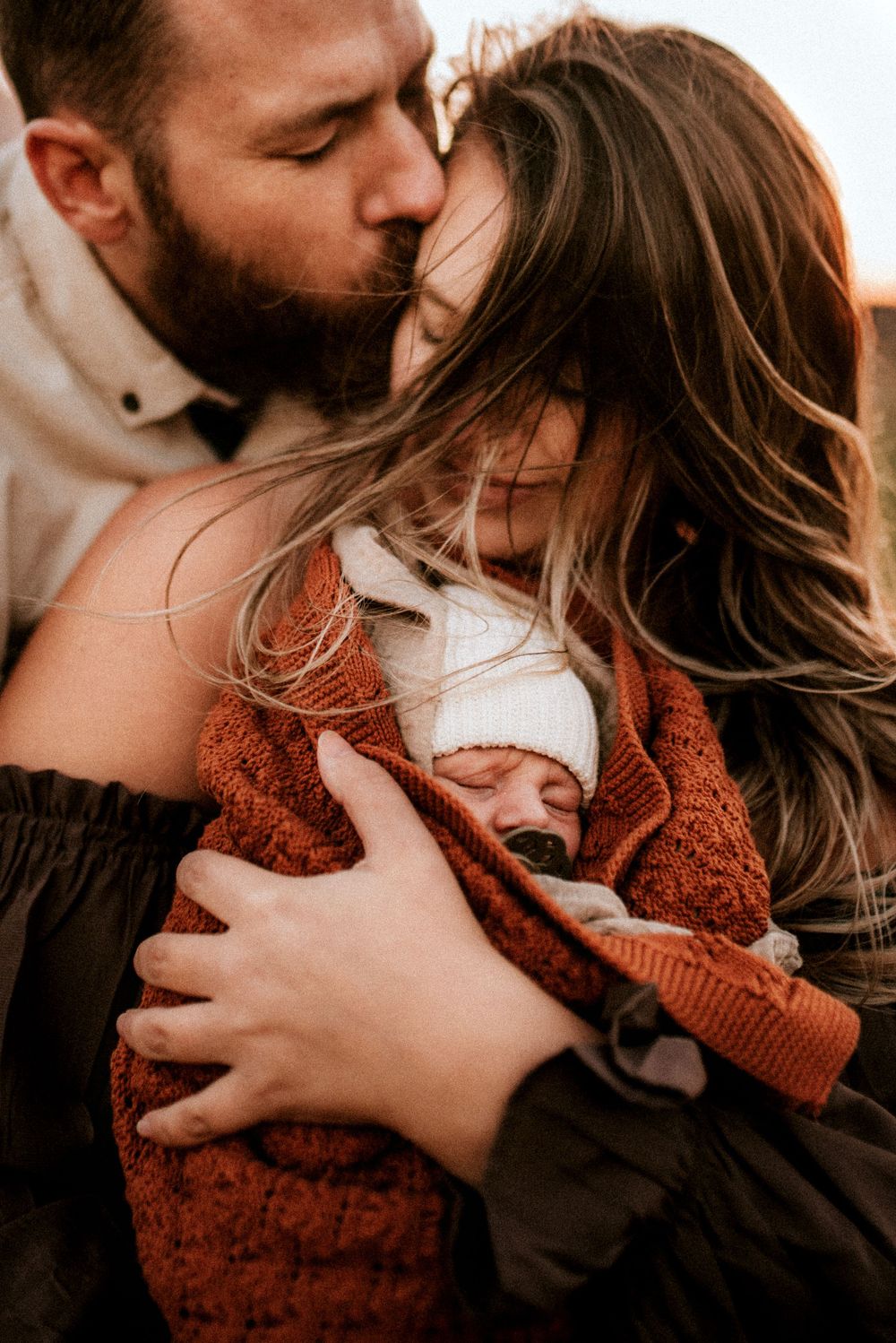 Image of Father kissing mother as her hair blows across her face in the wind and holding their newborn baby in the gorge of Oregon.