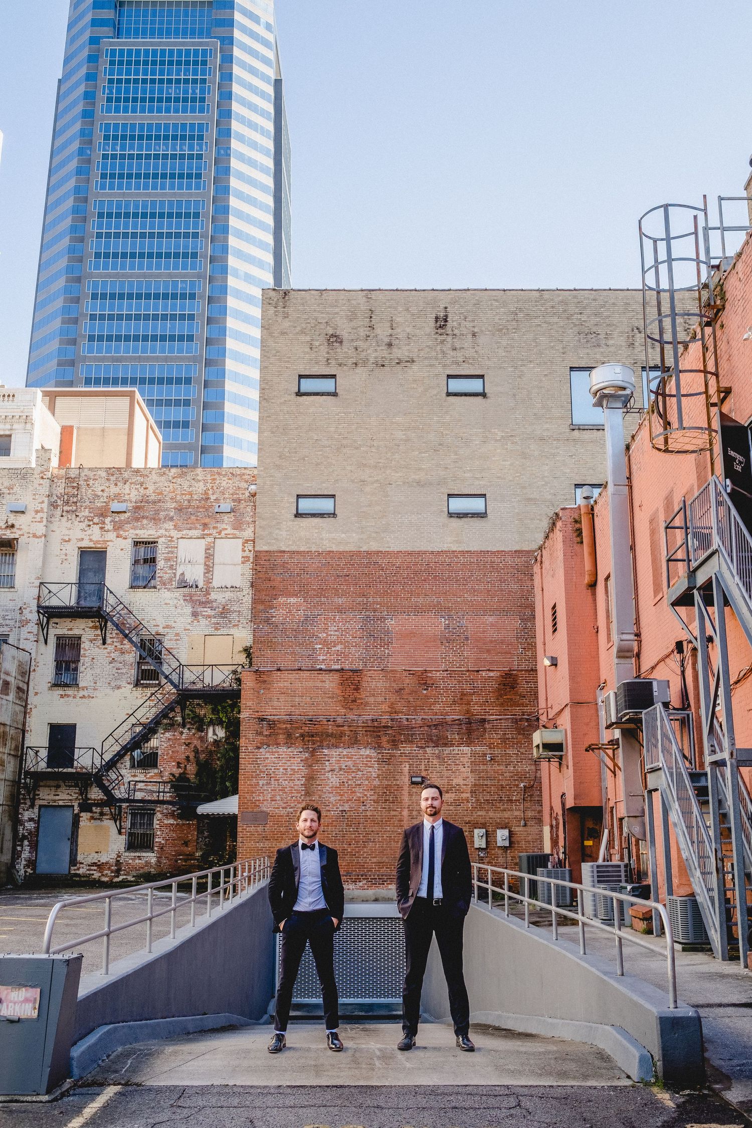 Two men in formal suits standing in an urban alley surrounded by brick buildings and a skyscraper.