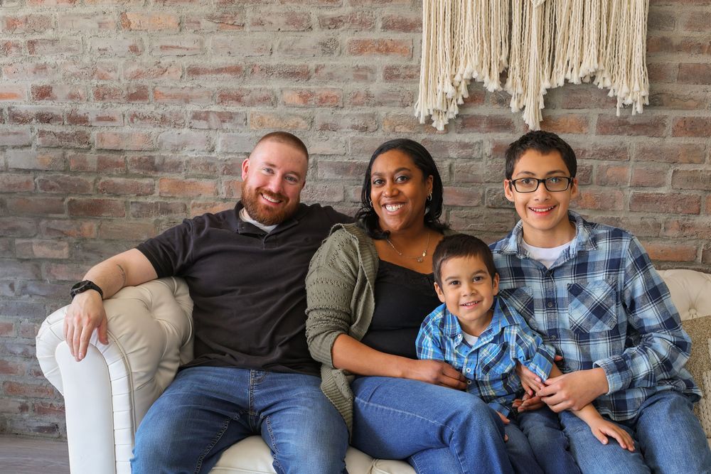 Family sits together on a gray couch against exposed brick wall with macrame wall hanging behind them.