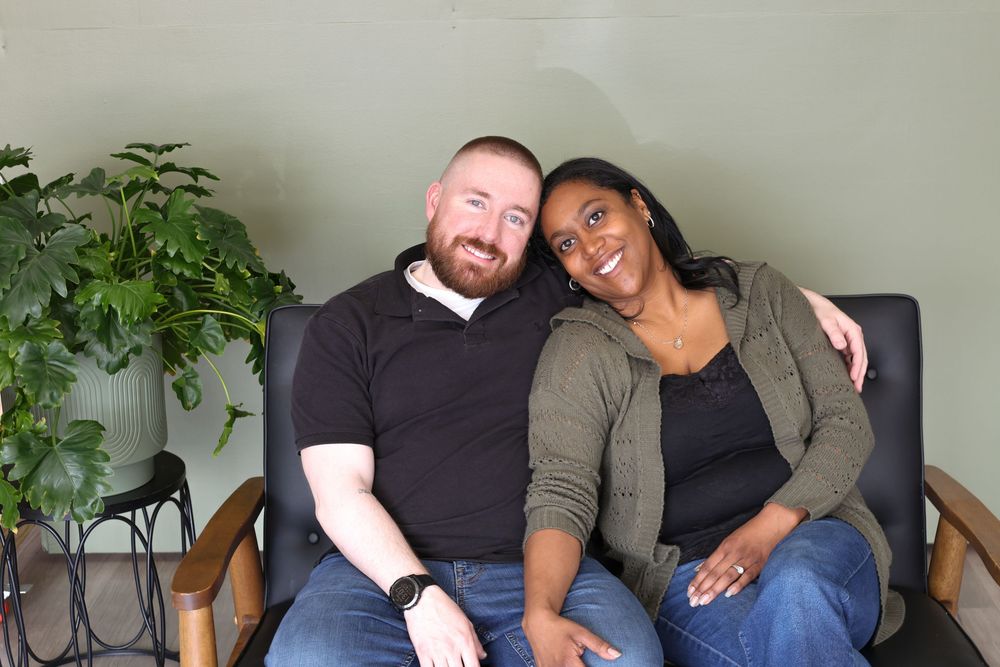 A couple sits close together on a wooden chair, with a green houseplant visible in the background.