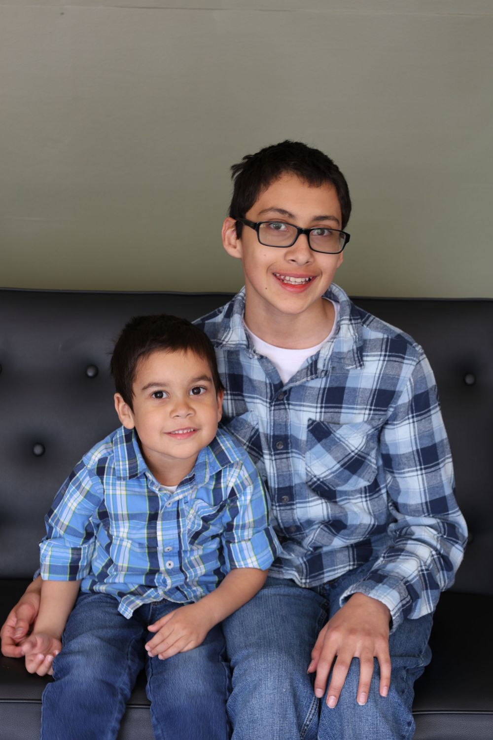 Two brothers wearing matching plaid shirts sit together on a black leather couch.