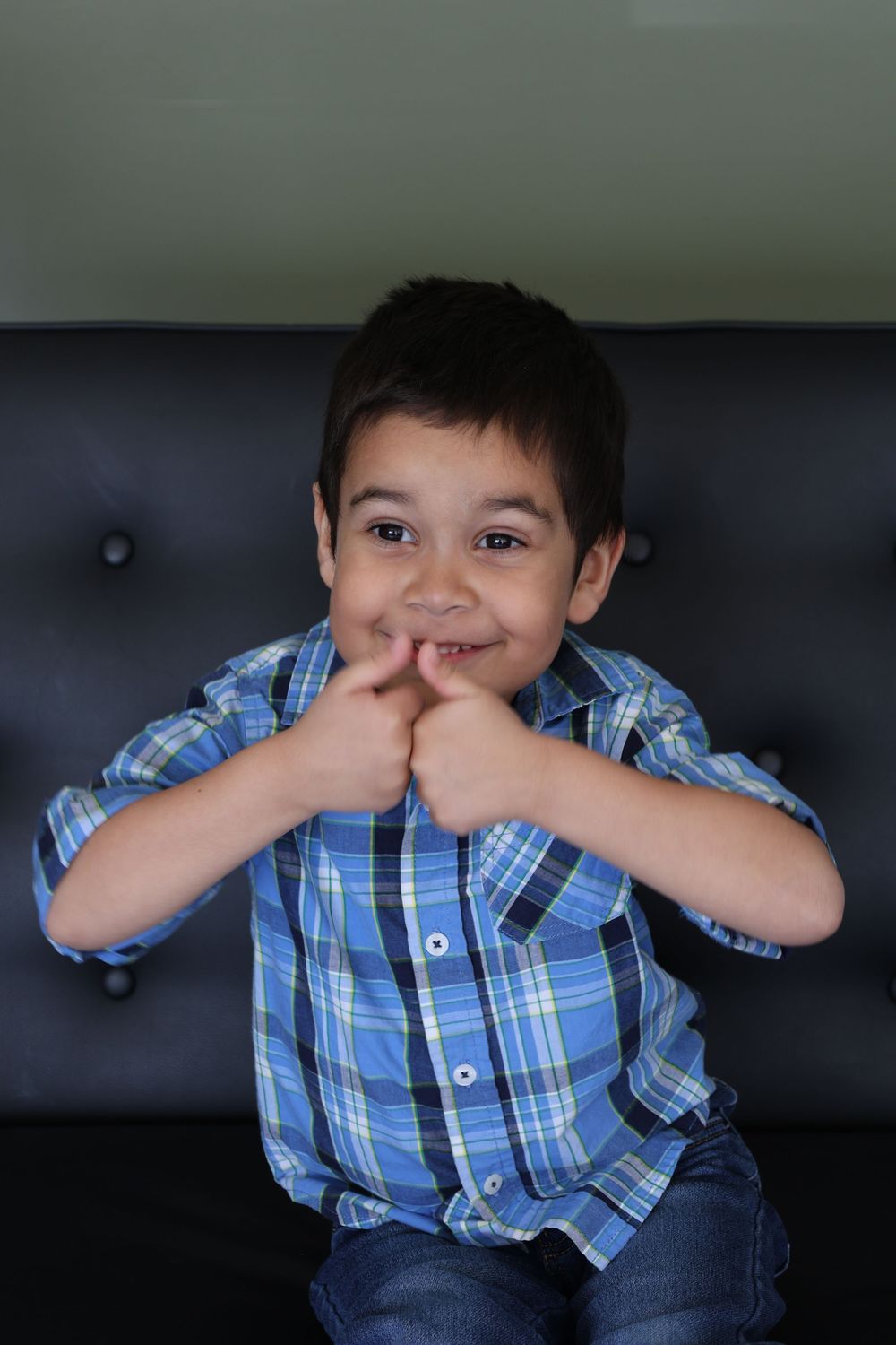 Child in blue plaid shirt sits on dark leather couch making silly faces and gestures.