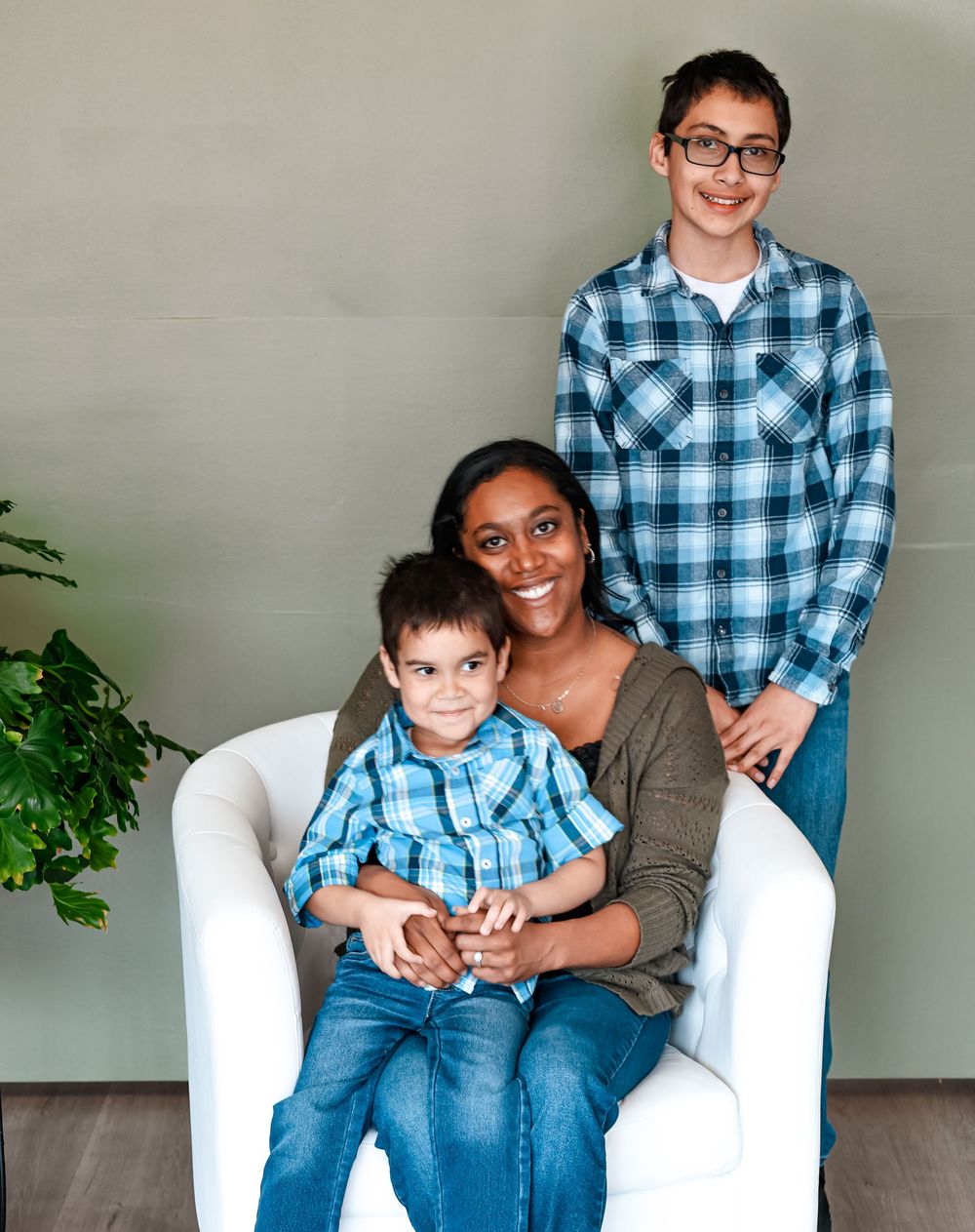 A family poses together in a living room, wearing coordinating blue plaid shirts and sitting on a white chair by a plant.